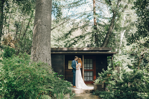 Bride and groom lovingly facing one another while standing next to a tree in front of a cabin.