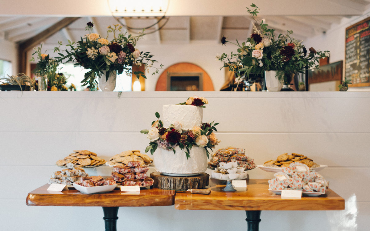 Table featuring white wedding cake adorned with floral confections along with cookies, pastries and other baked goods.